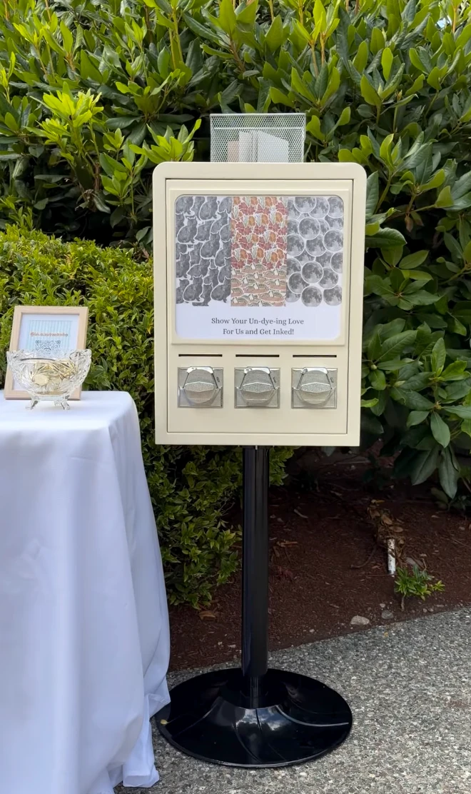 The vending machine on its stand at an outdoor wedding, with a sign reading 'Show Your Un-dye-ing Love For Us and Get Inked!' and a bowl of tokens nearby